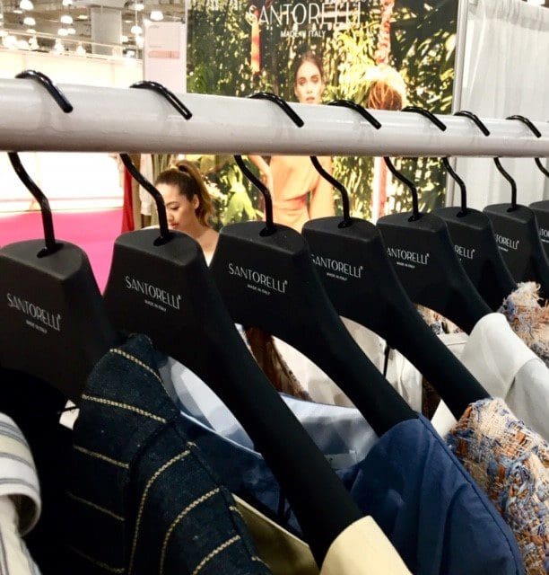 Display of Santorelli clothing on hangers at a fashion event, with a model visible in the background. Showcases Italian fashion design.