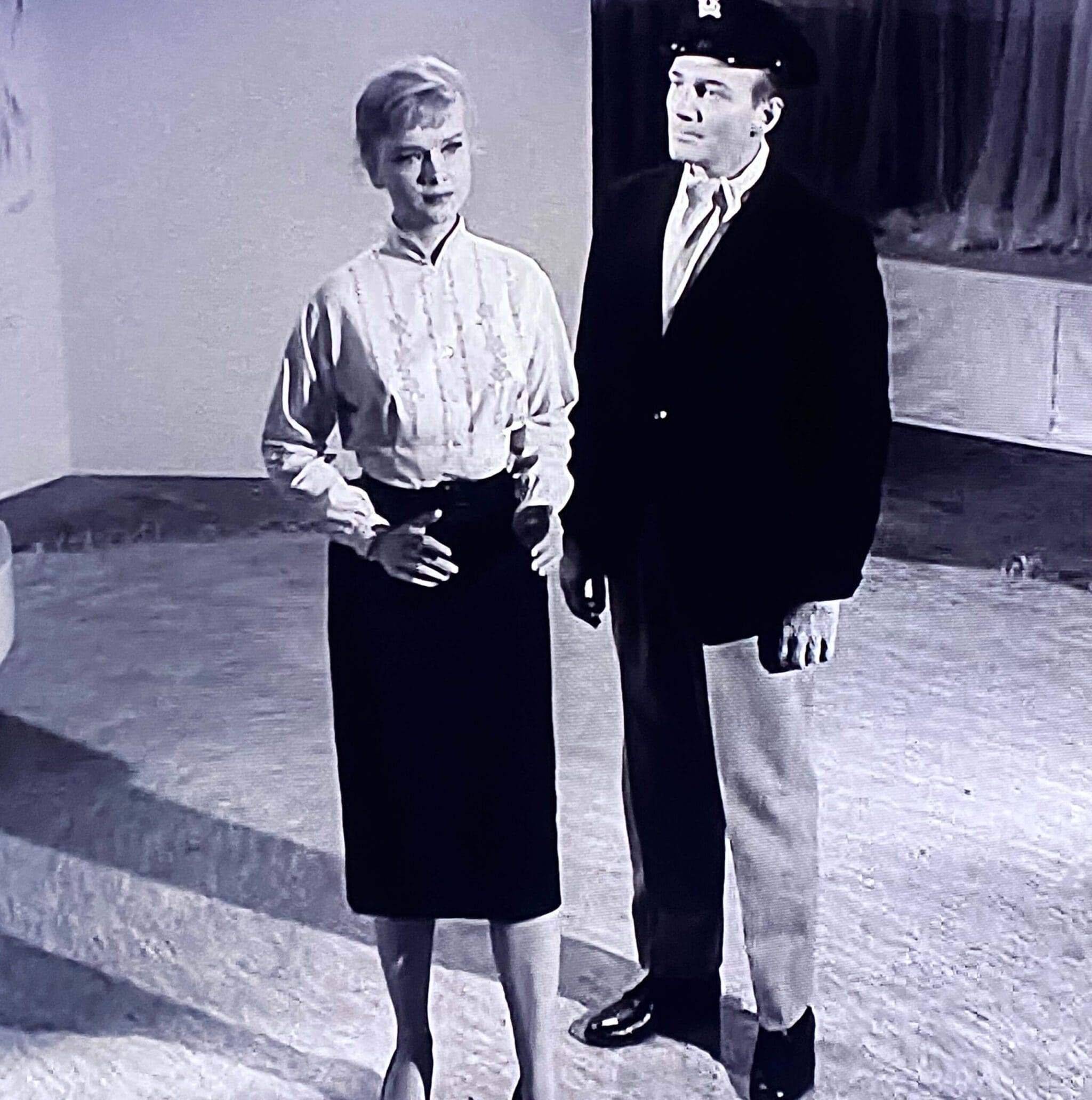 A woman in a blouse and skirt stands next to a man in a blazer and cap, both looking intently in a minimalist setting.