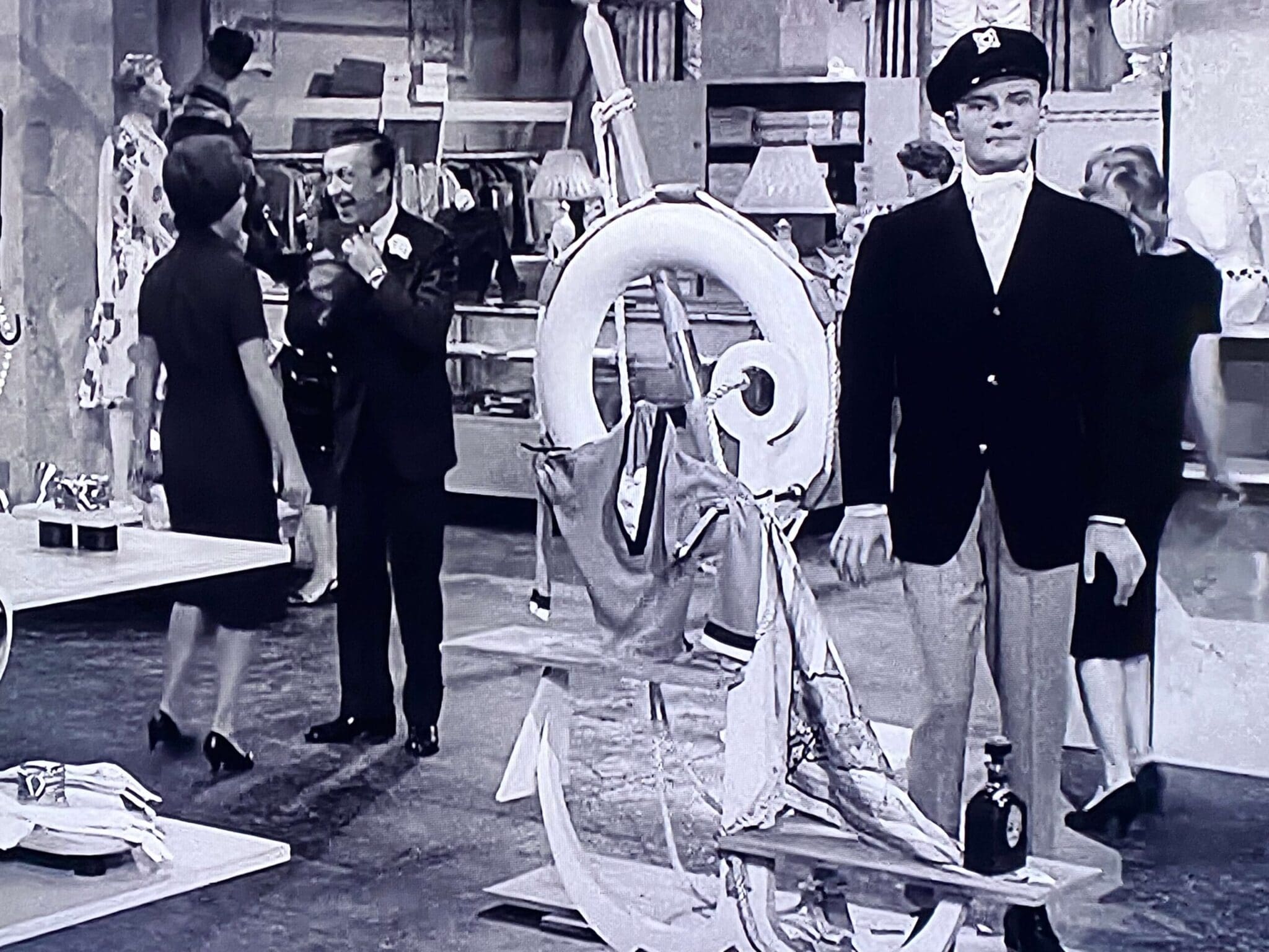 A dapper mannequin in a sailor outfit stands on display in a vintage clothing store, while a couple chats nearby, highlighting fashion in a retro setting.