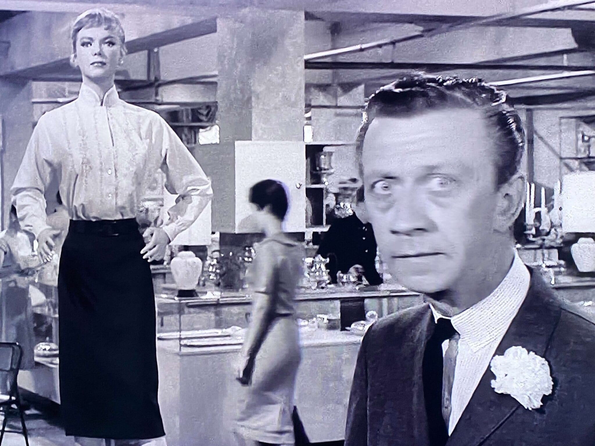 A tall mannequin in a blouse and skirt stands prominently in a bustling shop, while a man in a suit looks startled, enhancing the scene's comedic tension.