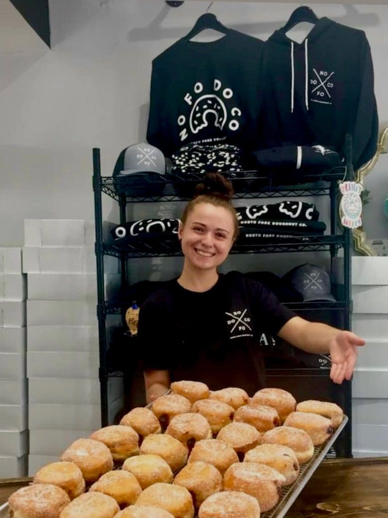 A smiling employee presents a tray of freshly made, powdered donuts in a bakery setting, with branded merchandise displayed in the background.