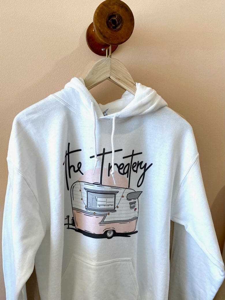 White hoodie featuring a graphic of a vintage food trailer and the text "The Treatery." Perfect for promoting a food-related brand or event.