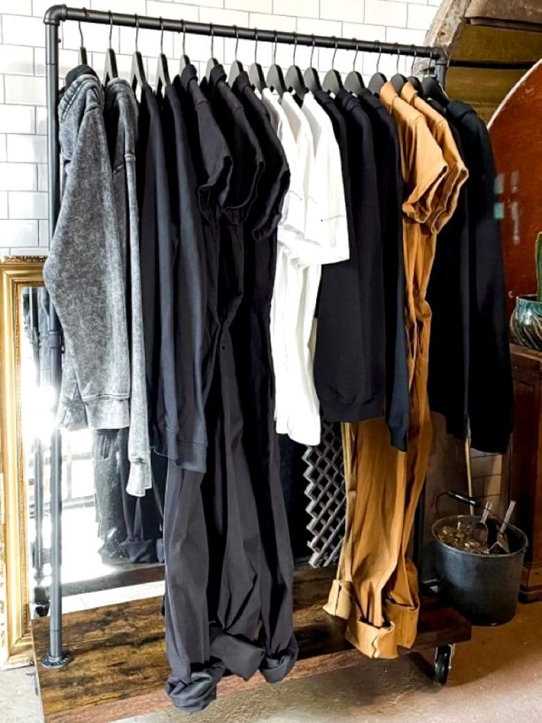A clothing rack displays a variety of casual garments, including hoodies, shirts, and pants in neutral tones, set against a rustic backdrop.