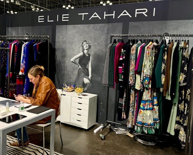 Elie Tahari booth with colorful garments on racks; stylist seated at table using phone.
