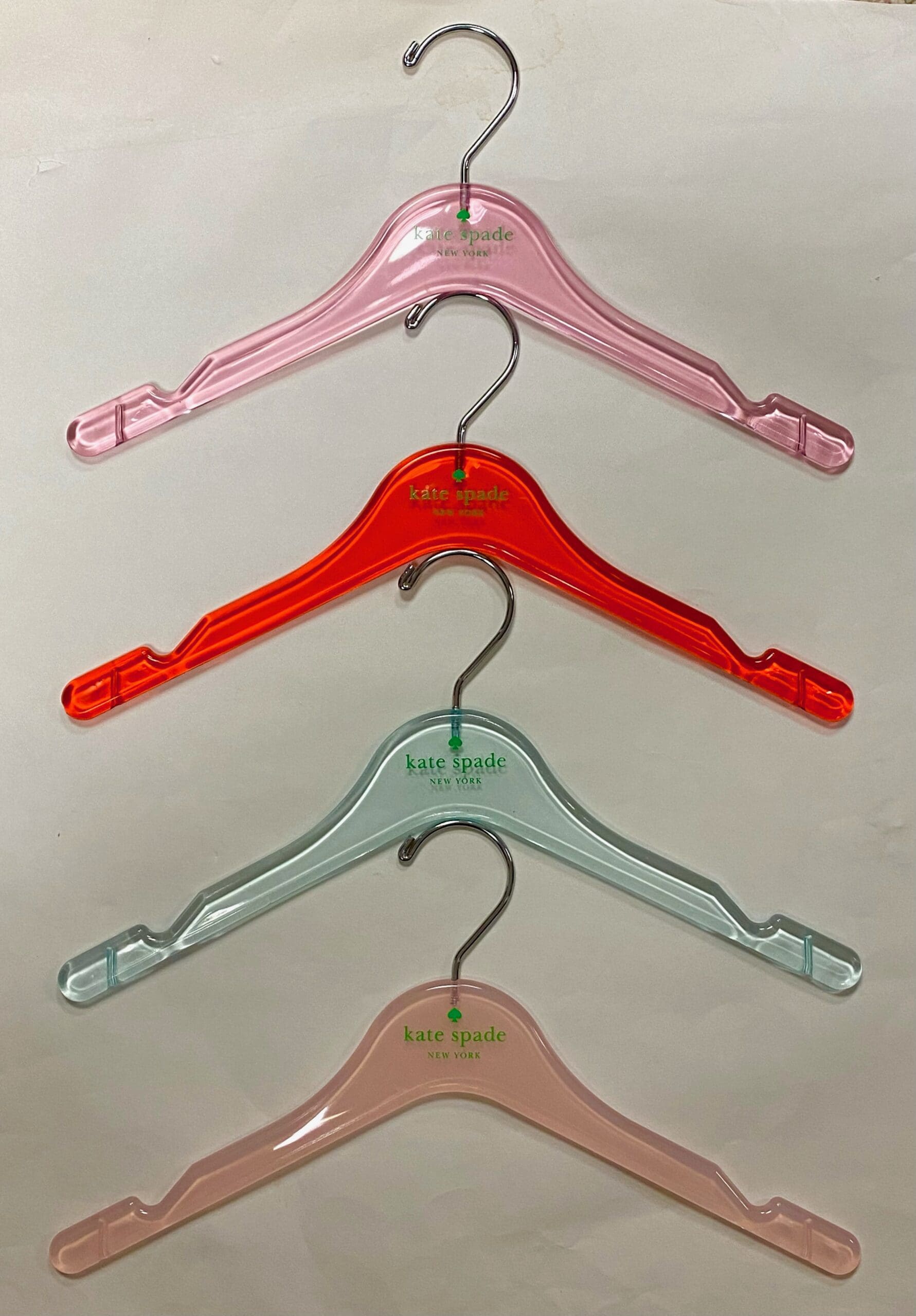 Four colorful plastic hangers by Kate Spade—pink, orange, light blue, and light pink—stacked horizontally against a light background.