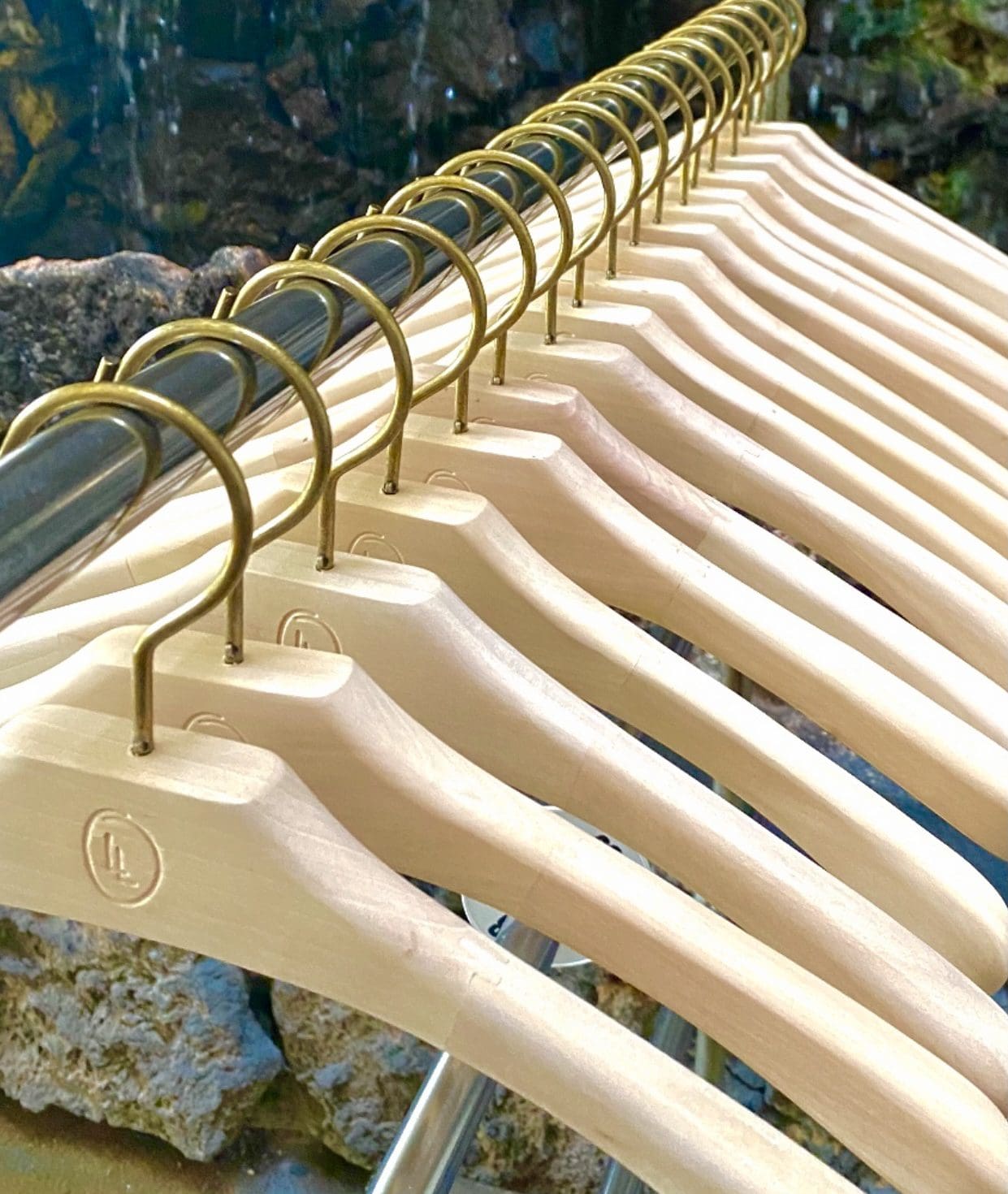 Wooden hangers with gold hooks are neatly arranged on a metal rack, set against a backdrop of a natural stone wall.