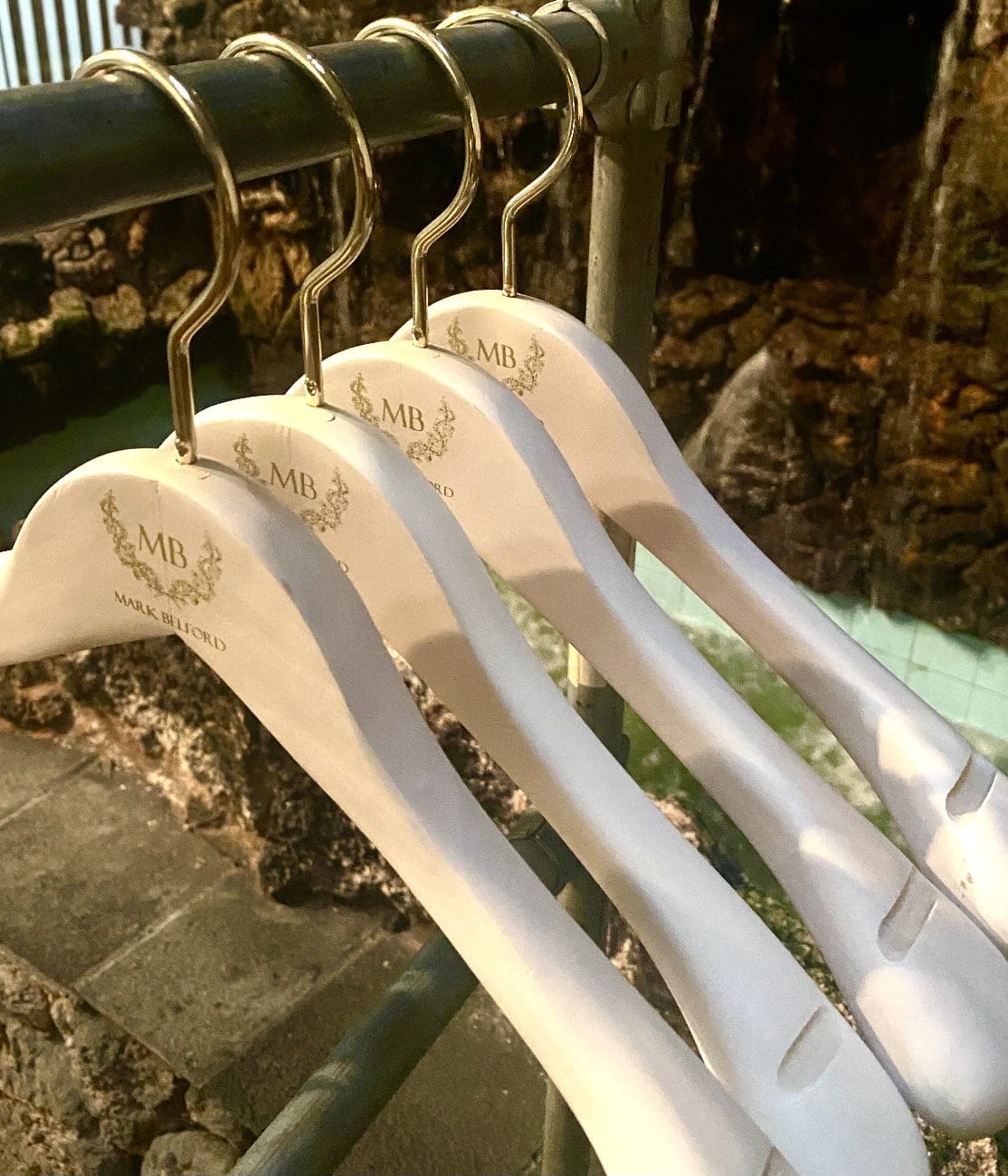 Four elegant, branded hangers hang on a metal rod, set against a backdrop of a serene water feature. Perfect for showcasing fashion items.