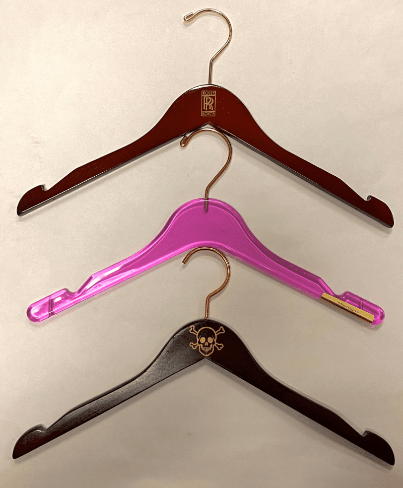 Three clothing hangers are displayed: a brown wooden hanger with a logo, a transparent pink plastic hanger, and a black hanger featuring a skull and crossbones.