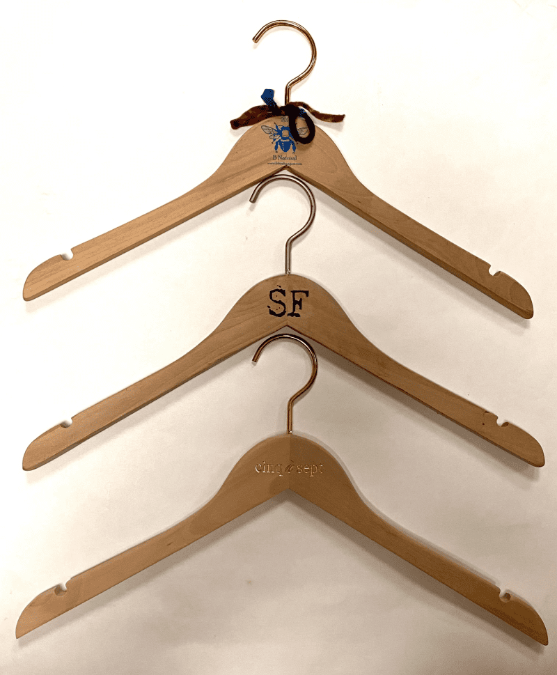 Three wooden clothing hangers arranged vertically. The top hanger features a bee logo, the middle is marked "SF," and the bottom reads "cinq/sept."