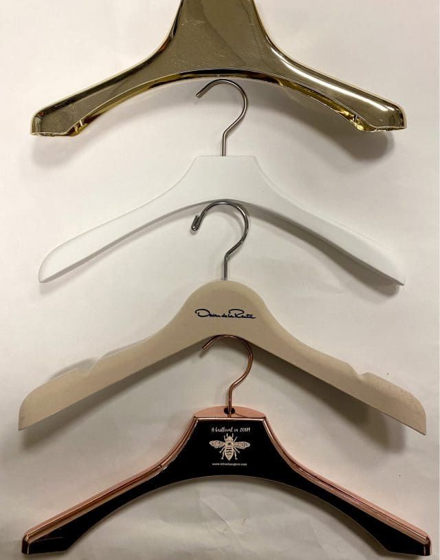 Four clothing hangers, featuring a shiny gold one, a plain white one, a beige hanger with a signature, and a black and rose gold design.