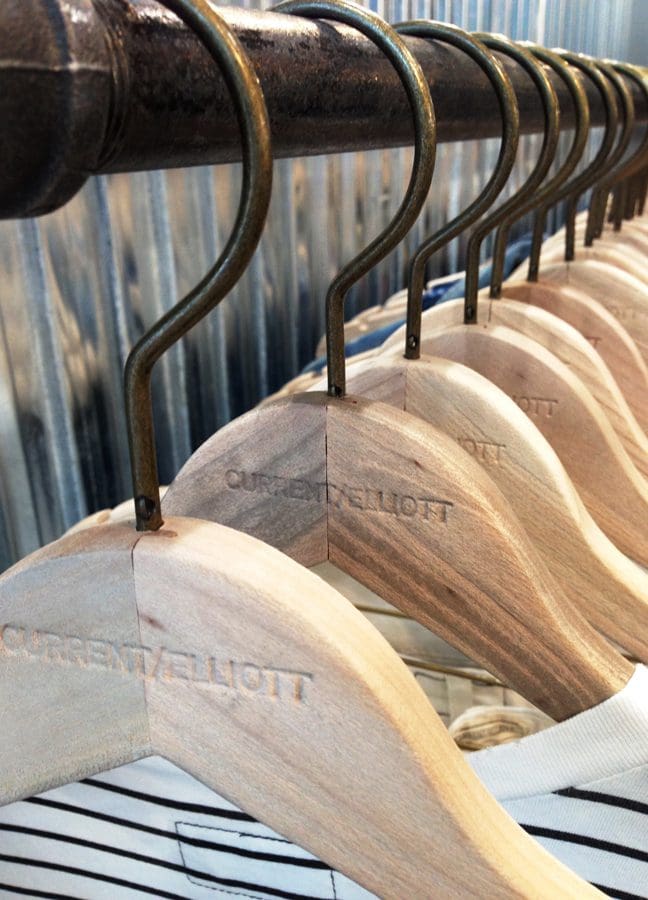 Wooden hangers with "CURRENT/ELLIOTT" branding showcase casual clothing in a boutique setting. The image highlights retail fashion presentation.